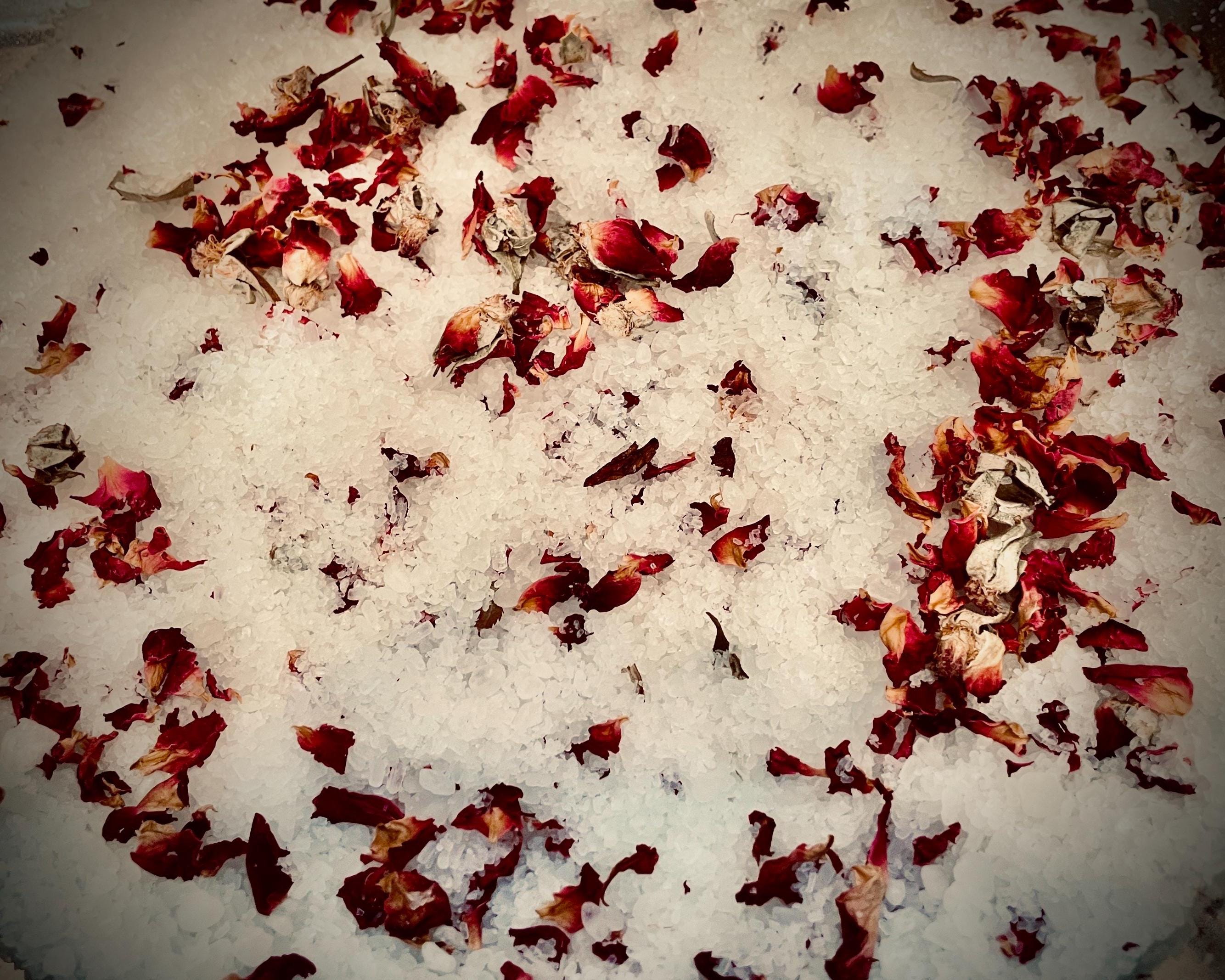 May include: Close-up of white bath salts with scattered red and dried rose petals. The image has a soft, diffused lighting, highlighting the texture of the salts and petals. The overall aesthetic is natural and spa-like.