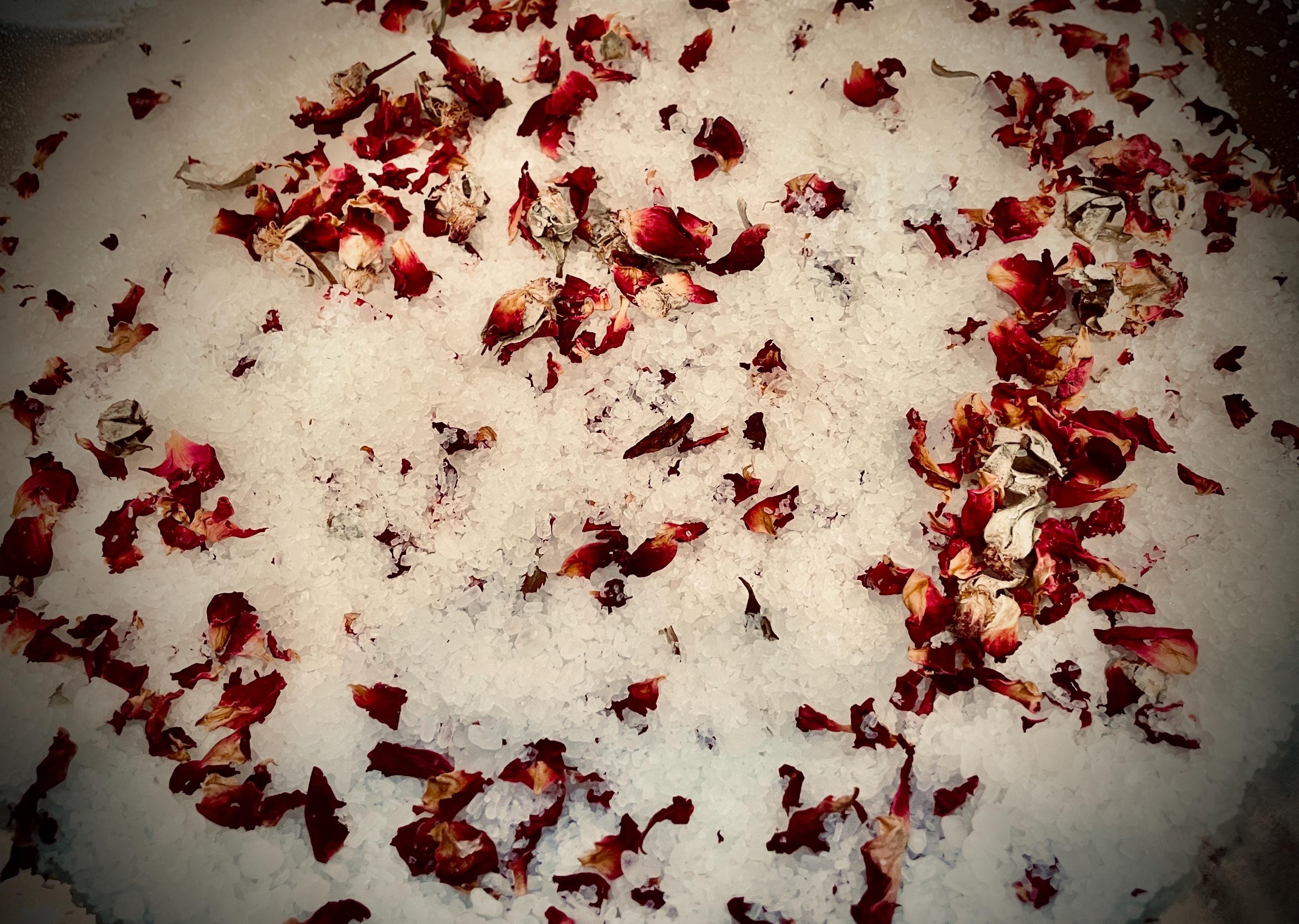 May include: Close-up of white bath salts scattered with dried red rose petals. The petals are various sizes and shapes, adding texture and color contrast to the image. The salts appear coarse and granular.
